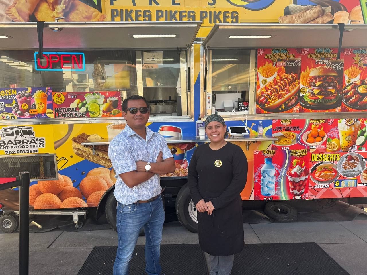 Two people smiling in front of a colorful food truck with menu items displayed.